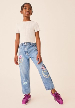 Pige iført en hvid tekstureret kortærmet top, lyseblå jeans med farverige lapper og lilla sneakers, stående mod en ensfarvet baggrund.
