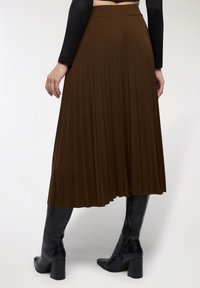 Gonna midi plissettata marrone con texture liscia, caratterizzata da vita alta e design fluido, abbinata a stivali neri con tacco al ginocchio.
