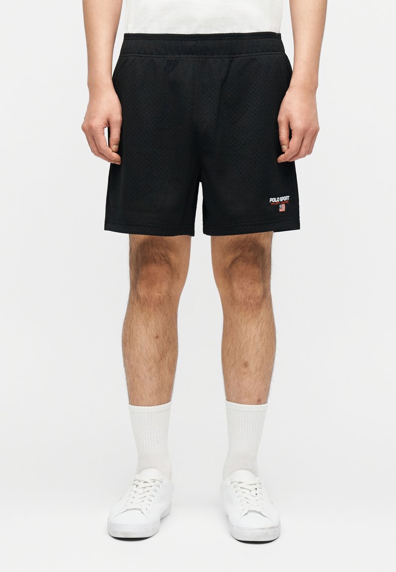 Άνδρας που φοράει μαύρα αθλητικά σορτς από δίχτυ με το λογότυπο "Polo Sport", λευκές κάλτσες και λευκά αθλητικά παπούτσια μπροστά σε απλό λευκό φόντο.