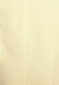 Vaaleankeltainen kangas, jossa on teksturoitu, poimutettu kuvio. Materiaali on sileää, hieman joustavaa ja tasaisesti taitettua. Ei näkyviä metalliosia tai koristeita.