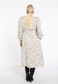 Vrouw met een lange blonde staart, gekleed in een witte midi-jurk met zwarte stippen, gouden oorhangers en zwarte haklaarzen, met haar rug naar de kijker.