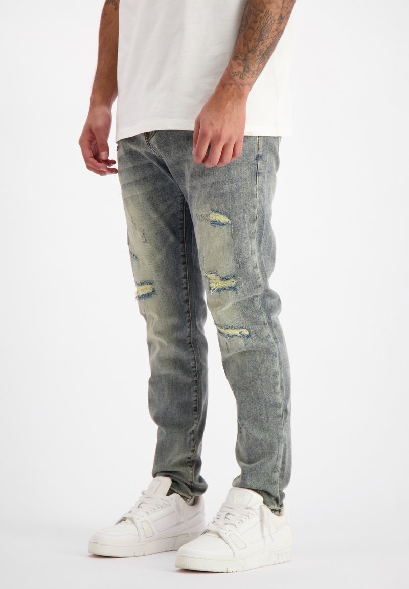 Vervaagde blauwe denim jeans met versleten gebieden en scheurpatronen. Slim fit ontwerp gecombineerd met witte sneakers.