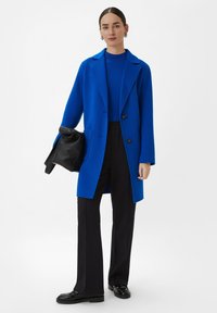 Blauwe lange jas met een gesneden revers, twee knopen en zijzakken, gecombineerd met een blauwe top en zwarte broek. Zwarte tas accentueert de look.