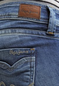 Vaqueros de mezclilla azul con una etiqueta de cuero con "Pepe Jeans" en relieve, costuras en contraste y un pequeño bolsillo bordado con el nombre de la marca.
