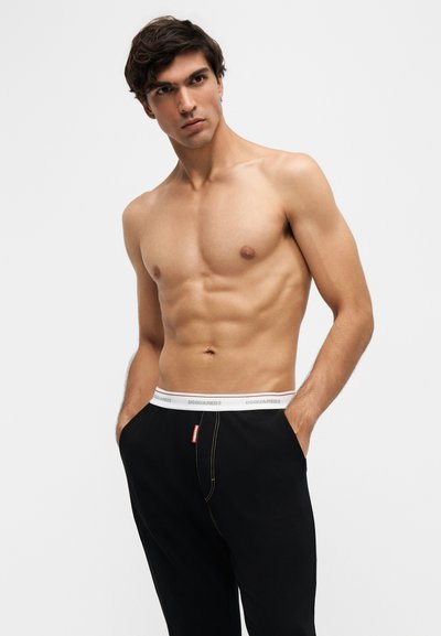 Jeune homme aux cheveux foncés posant torse nu, portant un pantalon noir avec une taille blanche sur un fond clair uni.