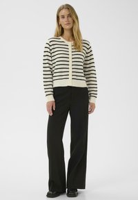 Femme aux longs cheveux blonds portant un pull boutonné blanc et noir à rayures, un pantalon noir à jambes larges et des chaussures noires, debout.