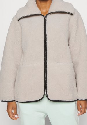 Personne portant une veste polaire gris clair avec fermeture éclair, col et poignets à bordure noire, associée à un pantalon de couleur claire.
