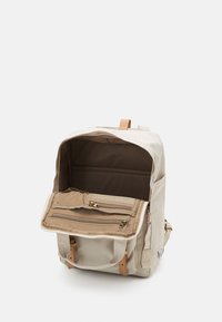 Beige backpack with a textured fabric, featuring a front zip pocket, leather accents, and an open main compartment.