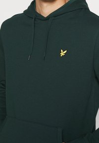 Felpa con cappuccio verde scuro realizzata in tessuto misto cotone, dotata di una tasca a marsupio e un piccolo logo di un uccello giallo ricamato sul petto.