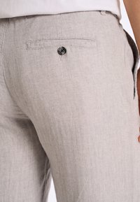 Pantalon gris clair en tissu chevron, avec une poche arrière unique dotée d'un bouton noir et des poches latérales.
