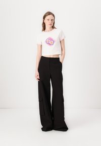 Valkoinen puuvillainen crop-top, jossa on vaaleanpunainen reunus ja värikäs sydängraafinen kuvio, yhdistettynä korkeavyötäröisiin mustiin leveälahkeisiin cargo-housuihin, joissa on taskut.