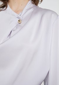 Gros plan sur un chemisier lavande clair avec un col drapé fermé par un seul bouton rond en or.