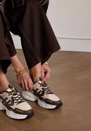 Beige en bruine sportschoenen met textuurpatronen, zwarte accenten en stevige zolen, die worden gestruikeld door een persoon in een donkere broek.