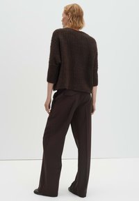 Brauner Pullover mit strukturierter Strickoptik und dreiviertel Ärmeln, kombiniert mit hoch taillierter, weiter brauner Hose. Schlichter Hintergrund.