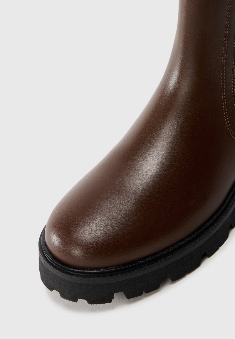 Brauner Lederstiefel mit schwarzer, dick gerippter Gummisohle vor weißem Hintergrund.