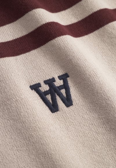 Tissu en tricot beige avec deux rayures horizontales rouge foncé et lettres noires "W" et "A" brodées en superposition.