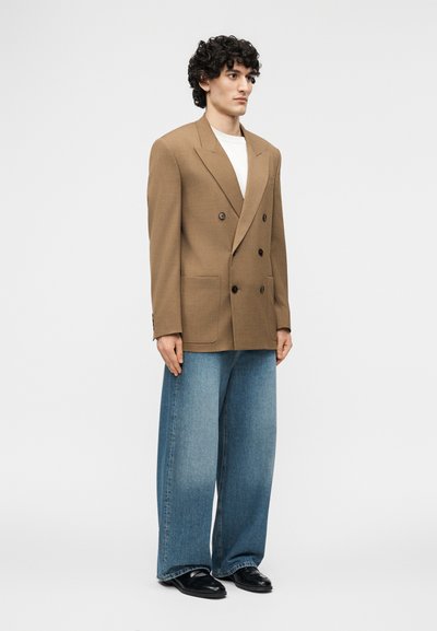 Άνδρας με σγουρά μαλλιά φορώντας καφέ διπλό πανωφόρι με κόπιτσες, λευκό πουκάμισο, φαρδιά μπλε τζιν και μαύρα παπούτσια, που στέκεται μπροστά σε απλό φόντο.