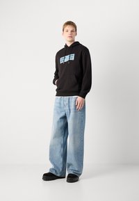 Zwarte hoodie met blauwe grafische tekst, gecombineerd met lichtblauwe wijde jeans en zwarte schoenen met een chunky zool.