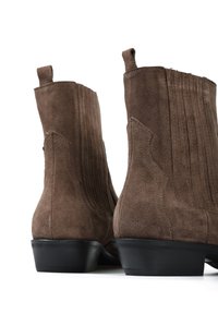 Brązowe zamszowe botki za kostkę z rowkowanym wzorem, stożkowatym kształtem i czarnymi gumowymi obcasami. Boczki z uchwytami ułatwiające zakładanie.
