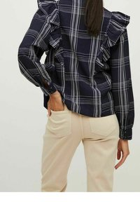 Navy plaid shirt with ruffled shoulder detail, long sleeves, and button cuffs; paired with beige pants. Fabrics appear smooth and lightweight.
