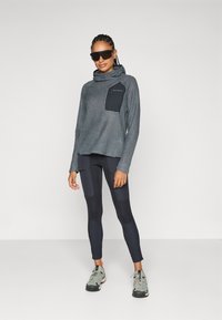 Haut de sweat à capuche texturé gris avec une poche poitrine noire, associé à des leggings sombres avec des détails en panneau et des chaussures de sport grises.