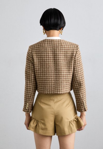 Blazer marrón de espiga con corte entallado, que presenta botones dorados y un cuello blanco, combinado con pantalones cortos beige con dobladillo con volantes.