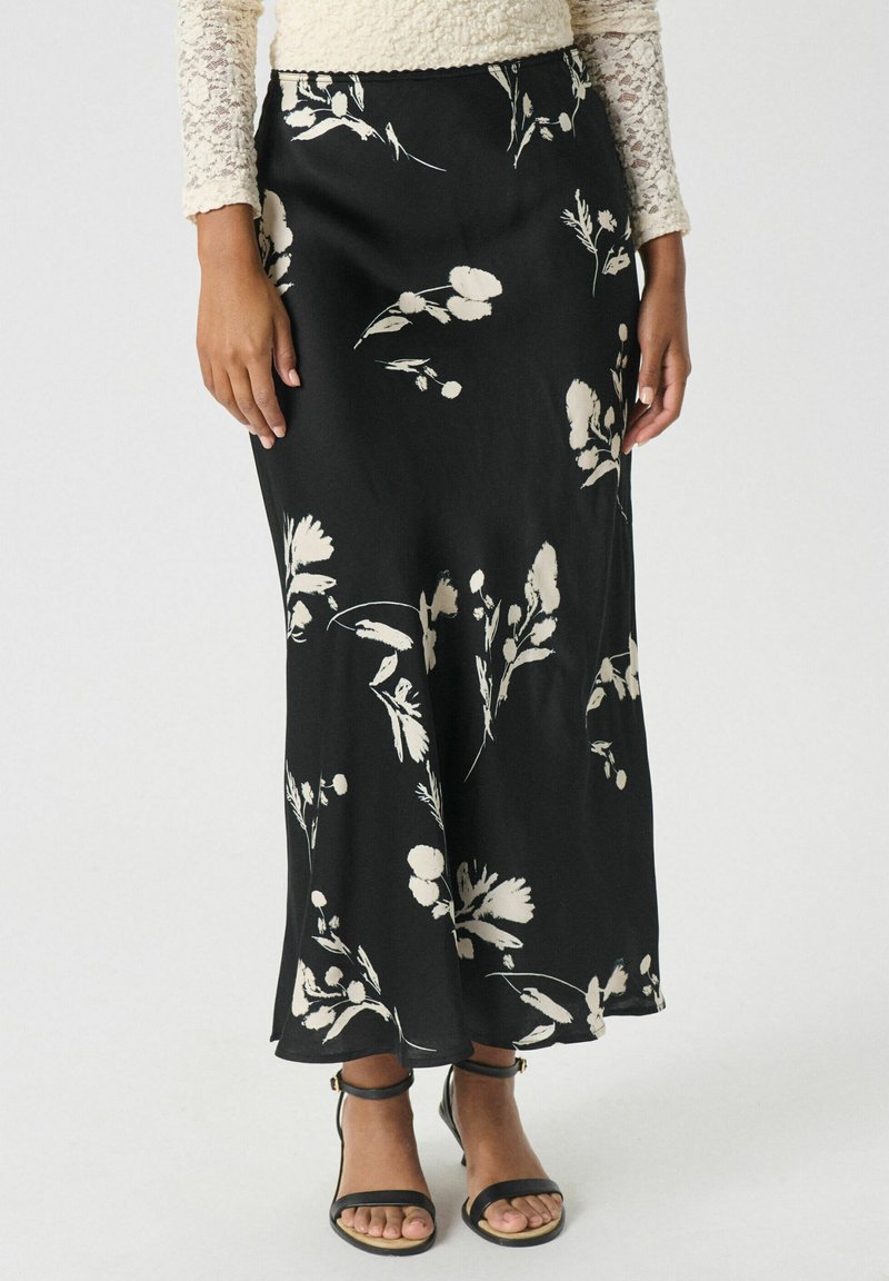 Jupe maxi noire à fleurs avec une finition satinée, ornée d'un imprimé botanique blanc. Texture lisse et silhouette droite. Sandales noires à brides.