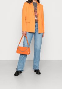 Blazer naranja con un diseño entallado, combinado con una camiseta gráfica, jeans azul claro y un pequeño bolso naranja con detalle de cadena.