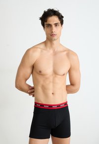 Calzoncillos boxer negros con una cinturilla elástica roja que presenta el nombre de la marca "HUGO" en texto blanco; tejido suave, diseño ajustado.