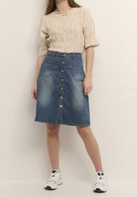 Ljusbeige, strukturerad, kortärmad topp i kombination med en mörkblå denimkjol med knappframsida och två sidofickor. Atletiska skor bärs.