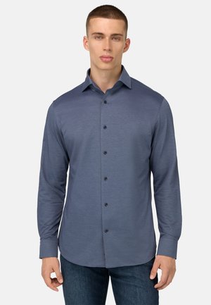 Langärmliges Hemd in mittlerem Blau, ausgestattet mit einem Standardkragen, schwarzen Knöpfen und einem abgerundeten Saum, kombiniert mit dunklen Denim-Jeans.