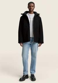 Juoda atspari orui striukė su gaubtu, užsegama užtrauktuku ir šoninėmis kišenėmis, dėvima ant šviesiai pilkos palaidinės ir mėlynų džinsų, su juodais batais.