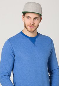 Blauwe sweatshirt met lange mouwen en een ronde hals, voorzien van contrasterende blauwe stiksels, gecombineerd met een lichtgrijze pet met een groene onderkant.