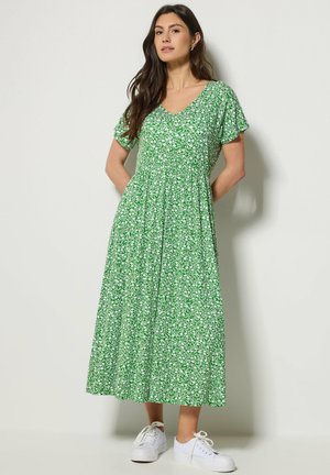 Vrouw met lang donker haar, gekleed in een groene midi-jurk met bloemenprint en witte sneakers, staand met handen achter de rug tegen een lichte muur.