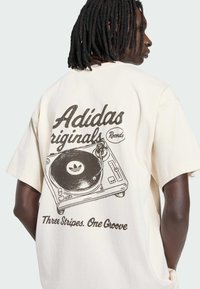 Bavlněné tričko krémové barvy s grafikou vintage gramofonu. Text zní "Adidas Originals Records" a "Three Stripes. One Groove."