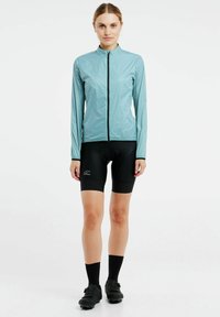 Lys blå, langærmet cykeljakke med lynlås; parret med sorte polstrede shorts og sorte cykelsko med spænder.