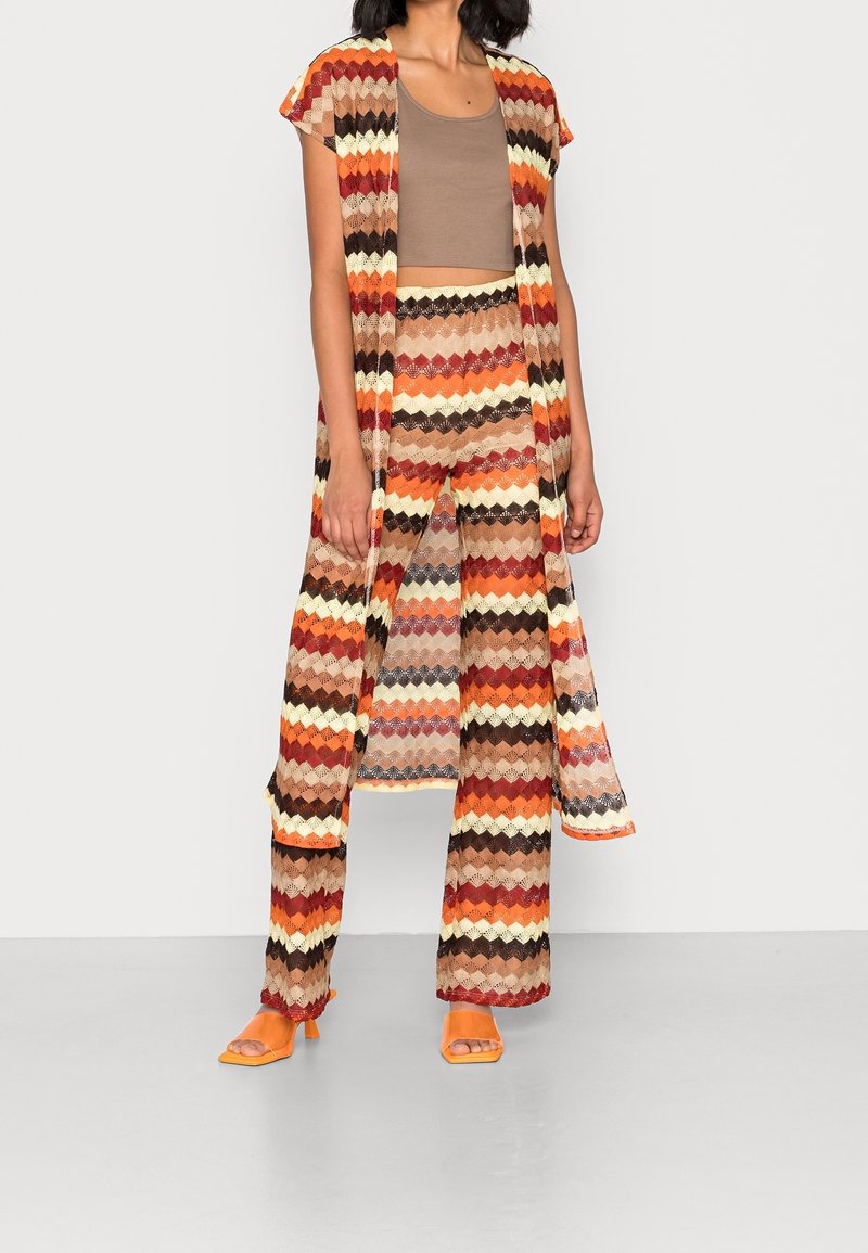 Knitted three-piece outfit featuring a striped, zigzag pattern in warm tones of orange, brown, and cream. Includes a sleeveless top, pants, and a long cardigan.