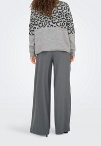 Femme aux cheveux bouclés portant un pull gris à motif léopard et un pantalon large gris, vue de dos sur un fond blanc.