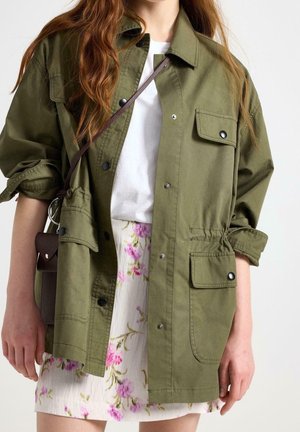 Femme portant une veste vert olive sur une chemise blanche et une jupe fleurie, portant un petit sac bandoulière marron foncé.