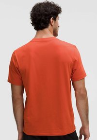 Bărbat cu păr creț și închis la culoare, purtând un tricou simplu portocalie cu mânecă scurtă, văzut din spate pe un fundal deschis.