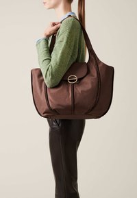 Brown textured handbag with a rounded shape, leather accents, and a gold circular hardware detail, carried by a person in a green sweater.