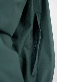 Gros plan sur la poche d'une veste verte foncée avec une fermeture éclair noire partiellement fermée, sur un fond clair uni.