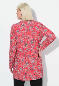 Punainen paisley-kuvioitu pitkähihaisuus tunika, jossa on pyöreä helma ja värikkäitä kukka- ja pyörteitä, yhdistettynä mustiin housuihin.