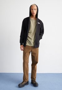 Czarna bluza z kapturem na zamek z logo, oliwkowa koszulka, brązowe spodnie sztruksowe i czarne buty na gumowej podeszwie; stojąc na jasnoniebieskiej powierzchni.