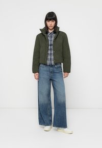 Olijfgroene, cropped jas van een gestructureerde stof met drukknoopjes, gedragen over een blauw ruitjeshemd. Gecombineerd met wijde denim jeans en witte sneakers.