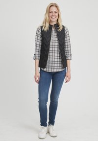 Čierny prešívaný vest s vysokým golierom na šedej a bielej kockovanej košeli. Modré džínsy a biele tenisky dotvárajú outfit.