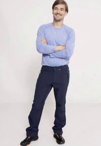 Sininen pitkähihaisen paidan ja tummansinisten ulkoiluhousujen kanssa, joissa on vetoketjutaskut. Malli seisoo käsivarret ristissä ja hänellä on mustat ja oranssit kengät.