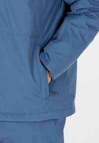 Blå, strukturerad ytterjacka med en ficka med dragkedja och kardborrefästen. Har en avslappnad passform och är tillverkad av lätt, ventilerande material.