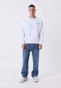 Sudadera blanca con estampado turquesa, jeans azules con estampado y zapatillas blancas. Apariencia general casual con un corte relajado y diseño básico.
