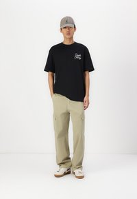 Sort bomuld T-shirt med et hvidt logo, parret med lysebeige cargo-bukser med flere lommer og en afslappet pasform, suppleret med en grå kasket.